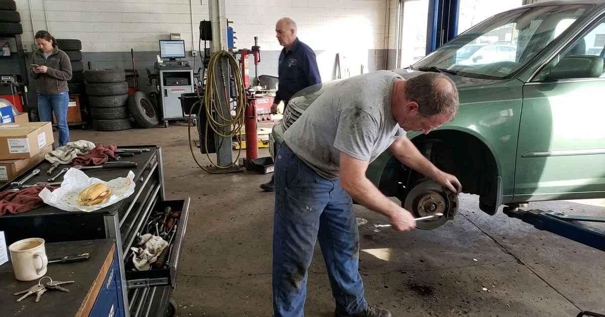 Technician checking steering and suspension components under a car, New Britain shop, candid